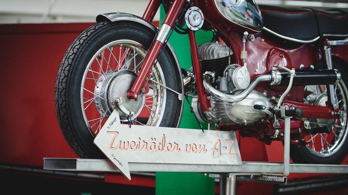 Eine rote Motorrad auf einem Aufsteller. Daneben befindet sich ein Schild mit der Aufschrift „Zweiräder von A-Z“. | © Königshofer