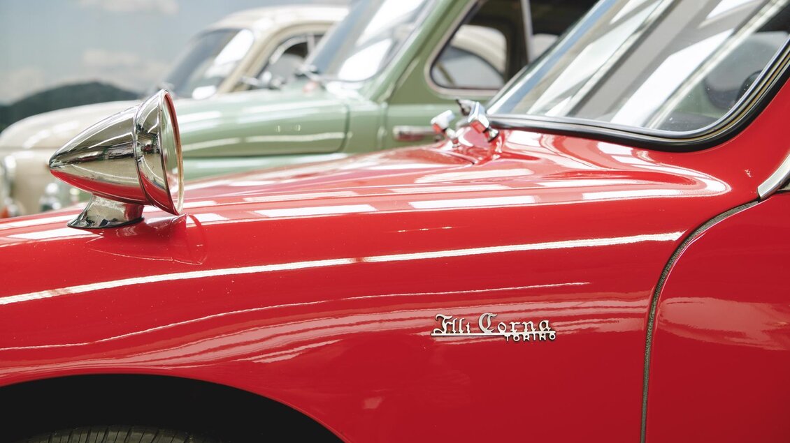 Ein rotes Auto mit glänzendem Scheinwerfer und einem klassischen Design. Im Hintergrund sind zwei weitere Fahrzeuge sichtbar. | © Königshofer