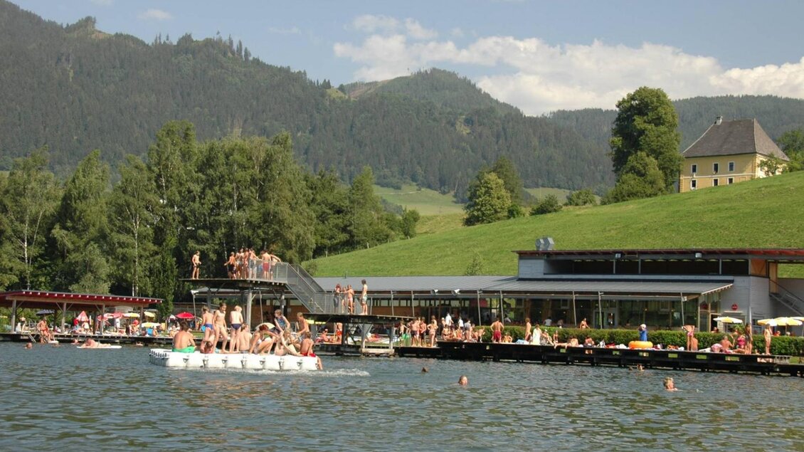 Ein schöner Badesee mit Menschen, die im Wasser schwimmen. Im Hintergrund sind grüne Hügel und ein Gebäude zu sehen.