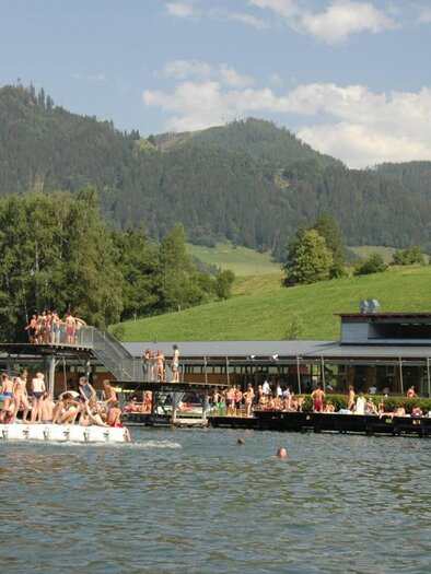 A beautiful bathing lake with people swimming in the water. In the background, green hills and a building can be seen.