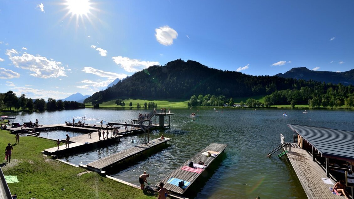 Ein sonniger Tag am See mit Badestränden und Menschen, die sich entspannen. Im Hintergrund sind grüne Wiesen und Berge zu sehen.