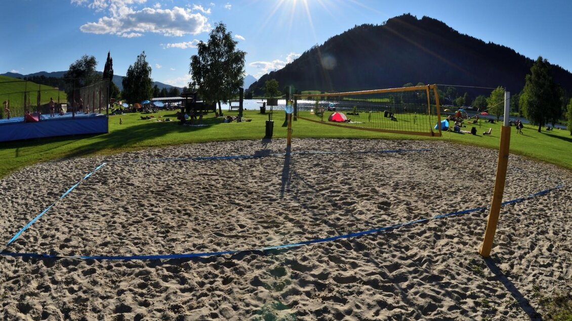 Ein Sandvolleyballfeld in einer sonnigen Umgebung, umgeben von Bäumen und Bergen. Im Hintergrund sind Menschen und Aktivitäten auf einer Wiese sichtbar. | © Putterersee