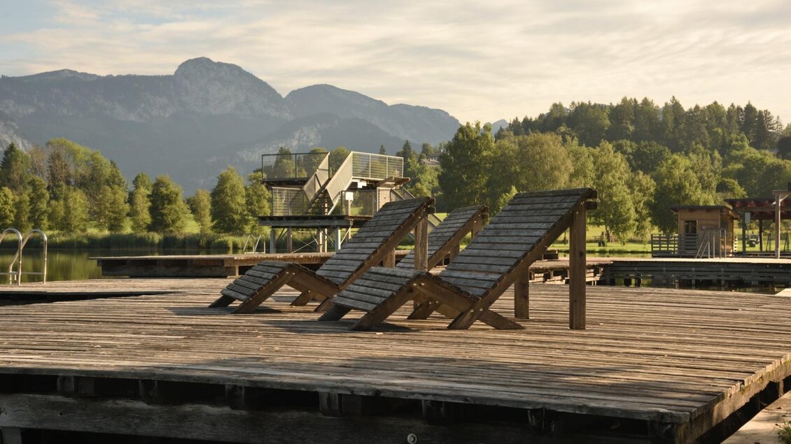 Eine Holzterrasse am See mit Liegen. Im Hintergrund sind Berge und Bäume zu sehen. | © Putterersee