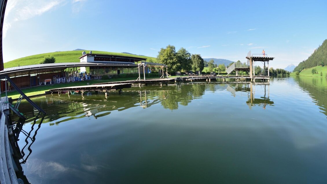 Ein klarer See mit Holzstegen und einer malerischen Landschaft im Hintergrund. Der Himmel ist blau und es gibt grüne Hügel um den See herum. | © Putterersee