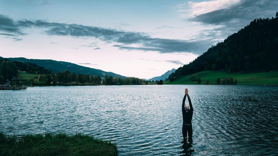 Ein Mensch steht im Wasser eines ruhigen Sees und streckt die Arme in die Höhe. Im Hintergrund sind grüne Wiesen und bewaldete Berge unter einem bewölkten Himmel zu sehen. | © Armin Walcher