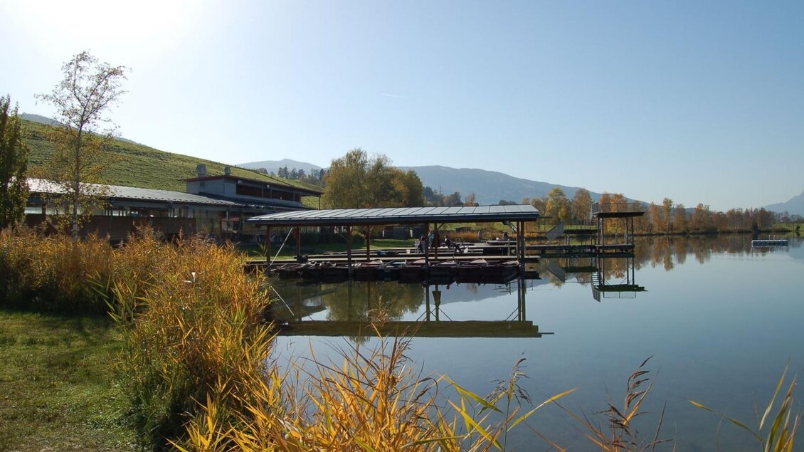 Ein ruhiger See mit Holzstegen und umgebenden Bäumen. Die Sonne scheint klar über die Landschaft. | © Putterersee