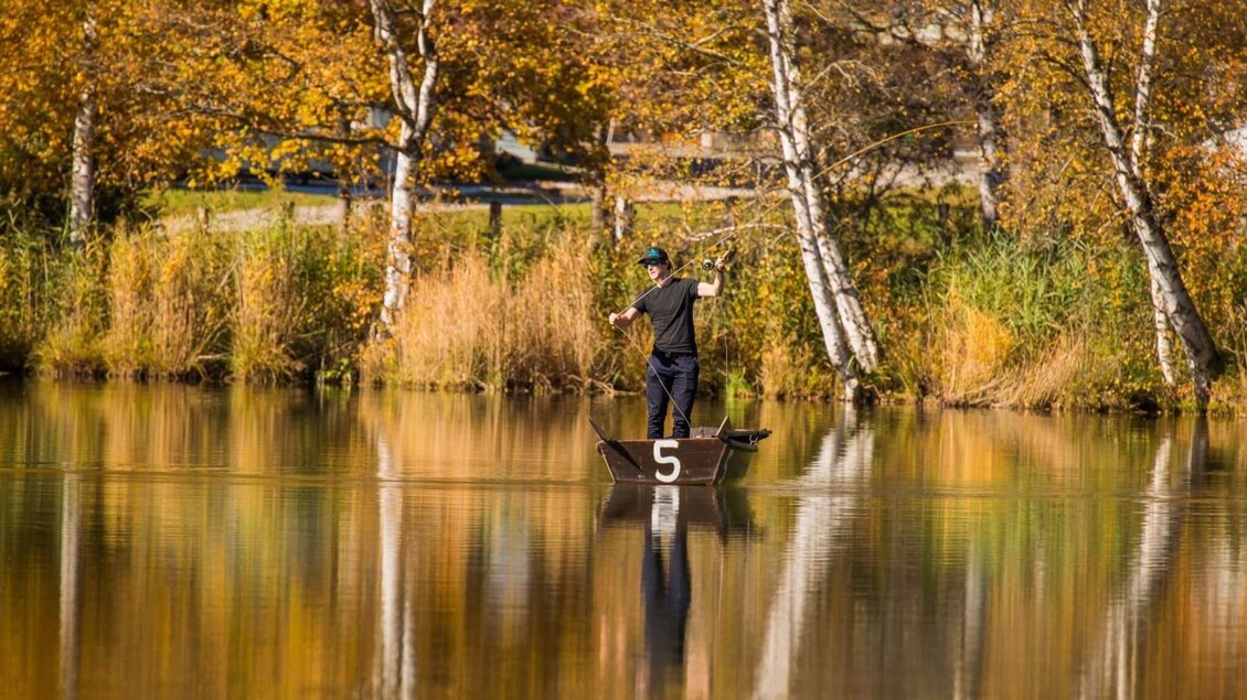 Ein Mann steht in einem Boot auf einem ruhigen See, umgeben von herbstlichen Bäumen. Das Wasser spiegelt die Farben der Umgebung wider. | © Putterersee