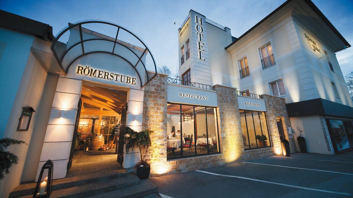 Das Bild zeigt den Eingang eines Hotels mit dem Schriftzug "Römerstube". Die Fassade ist beleuchtet und hat eine moderne Architektur. | © Römerstube