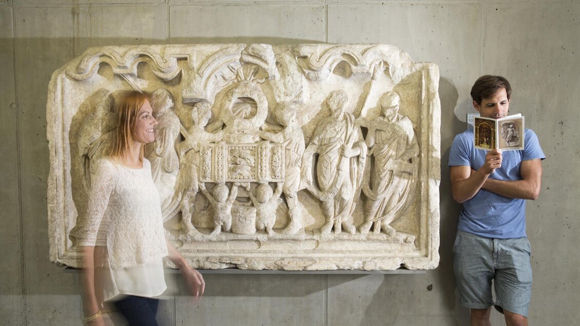 Eine historische Reliefdarstellung ist an der Wand zu sehen. Zwei Personen stehen in der Nähe; eine Frau geht vorbei, während ein Mann ein Buch betrachtet. | © Bernhard Bergmann