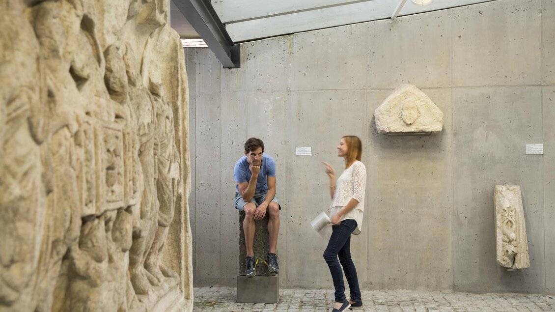 Eine Person sitzt auf einem Block und hört einer anderen Person zu, die im Gespräch ist. Im Hintergrund sind alte Steinskulpturen und eine moderne Architektur zu sehen. | © Bernhard Bergmann