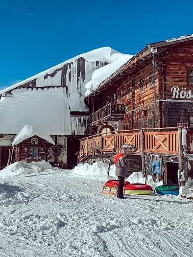 A rustic mountain cabin surrounded by snow. In the foreground stands a person with a sled. | © Oldenburg, Christin