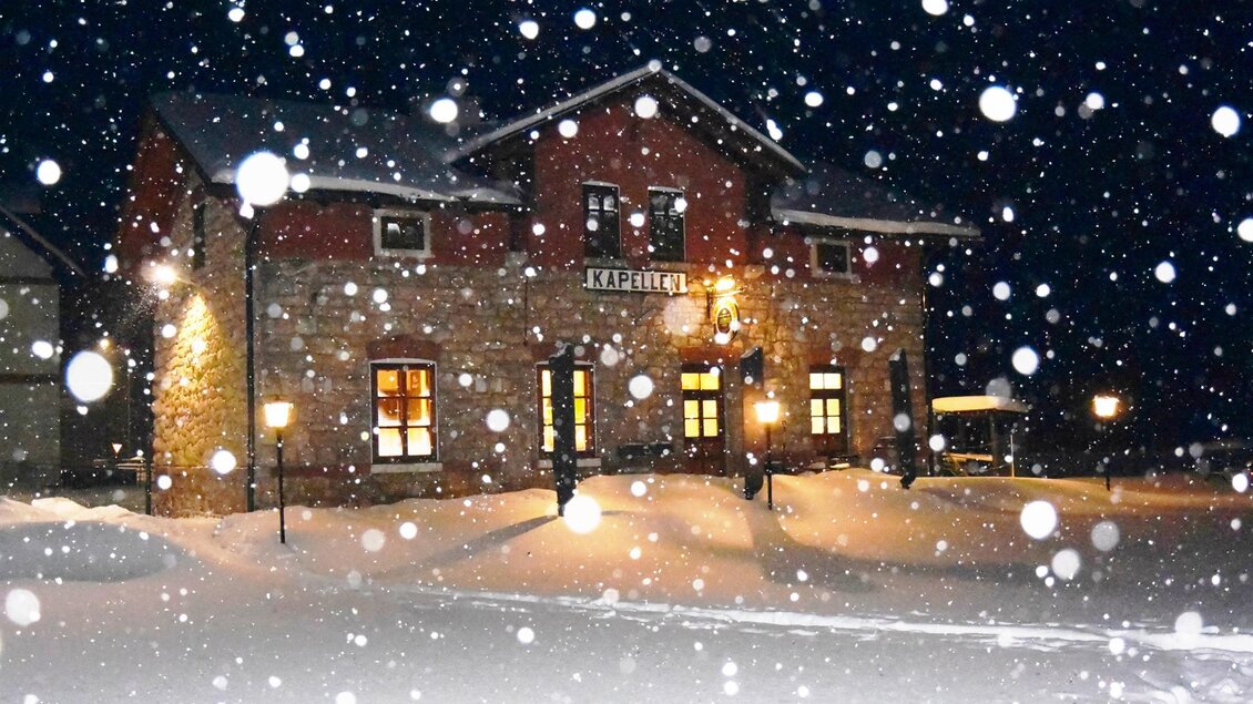 Ein charmantes Haus im Schnee, umgeben von winterlicher Atmosphäre. Große Schneeflocken fallen sanft über die Szenerie in der Nacht. | © Röststation