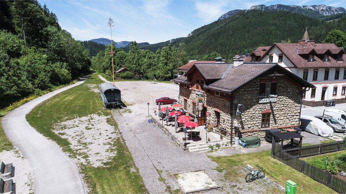Ein altes Gebäude mit einem Steinfassade und roten Sonnenschirmen. Im Hintergrund sind grüne Wälder und Berge zu sehen. | © Röststation