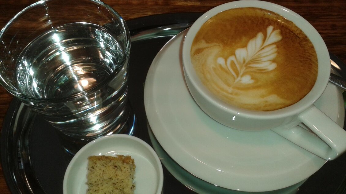 Ein schöner Kaffee mit einem kunstvollen Milchschaummuster in einer weißen Tasse. Dazu ein Glas Wasser und ein kleines Stück Gebäck auf einem Etagere. | © Röststation