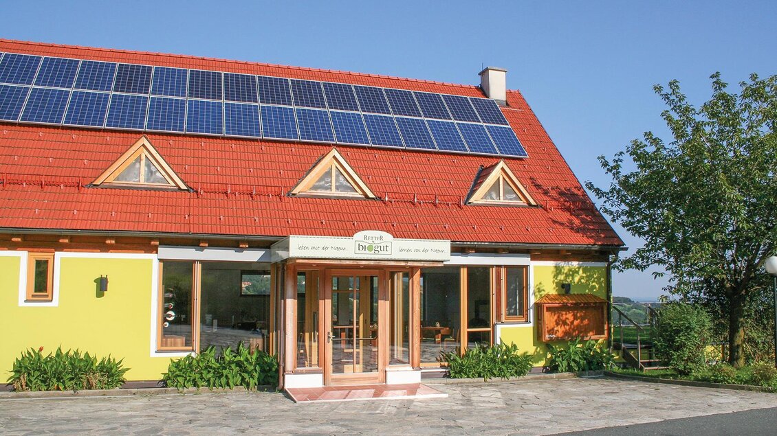 Ein modernes Haus mit einem rot gedeckten Dach und Solarpanelen. Es hat eine gelbe Fassade und ist von grünen Pflanzen umgeben. | © Helmut Schweighofer