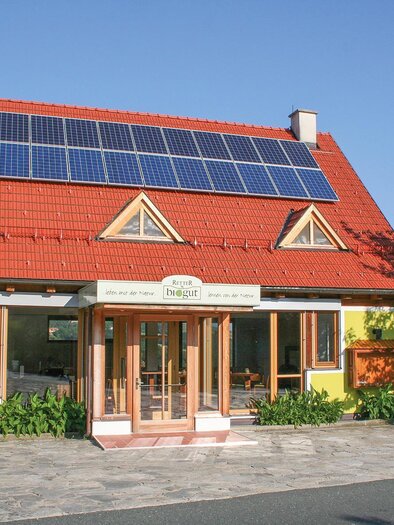 Egy modern ház vörös tetővel és napelemekkel. Sárga homlokzata van, és zöld növények veszik körül. | © Helmut Schweighofer