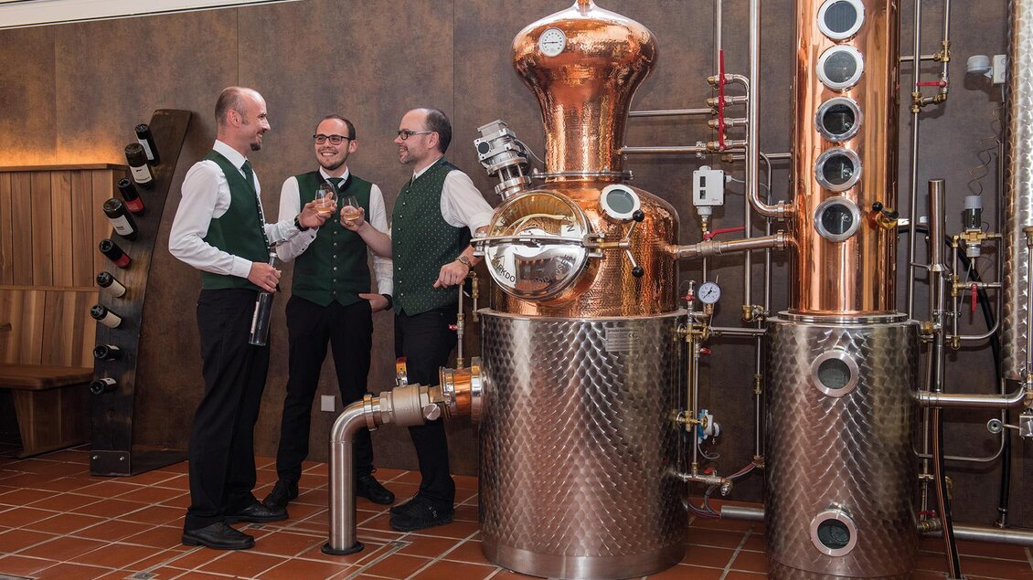 Drei Männer in eleganter Kleidung stehen neben einer Kupfer-Distillerie. Sie sprechen und halten Getränke in einer rustikalen Umgebung. | © Helmut Schweighofer