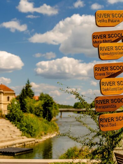 A beautiful river with a sign pointing to various places. Above the water, some clouds can be seen in the sky. | © Thermen- & Vulkanland