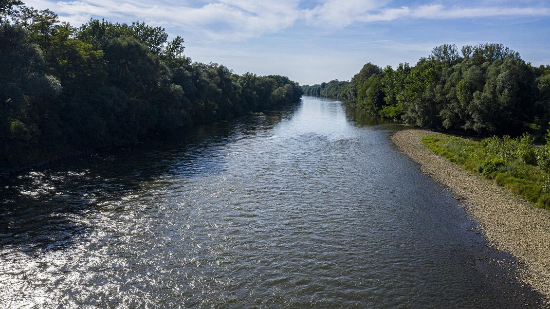 Ein ruhiger Fluss fliesst durch eine grüne Landschaft. Am Ufer sind Bäume und ein Kiesstrand sichtbar. | © Thermen- & Vulkanland