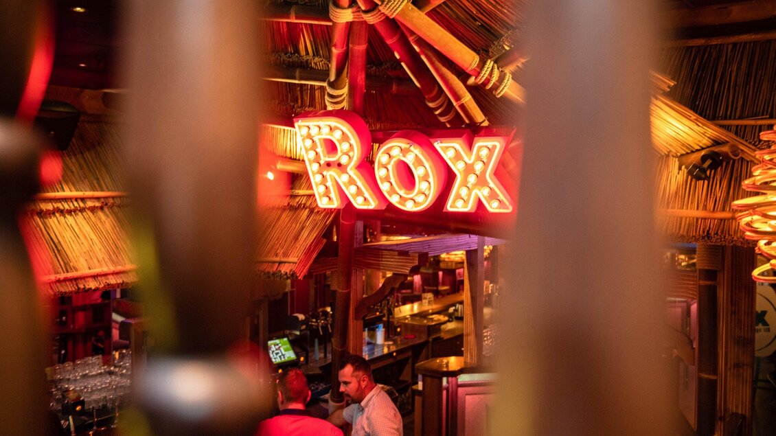 Ein gemütlicher Innenraum mit einer neonbeleuchteten Bar namens "Rox". Die Atmosphäre ist einladend und tropisch gestaltet. | © ROX - Kyrillos Mikhail