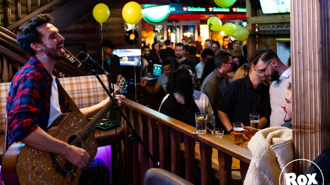  Sänger mit Gitarre tritt vor Publikum in einer vollen Bar auf, Gäste unterhalten sich und trinken Bier. | © ROX