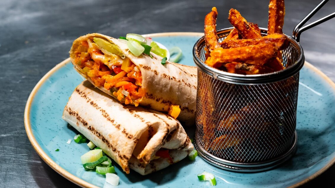 Halbierter Wrap mit Gemüse und Käse, garniert mit Frühlingszwiebeln, dazu Süßkartoffel-Pommes im Metallsieb. | © ROX