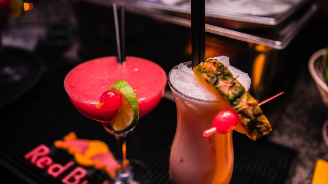 Zwei bunte Cocktails stehen auf der Bar. Der erste ist rot mit einer Limettenscheibe, der zweite ist hell mit einer Ananasscheibe und einer Kirsche. | © ROX - Kyrillos Mikhail