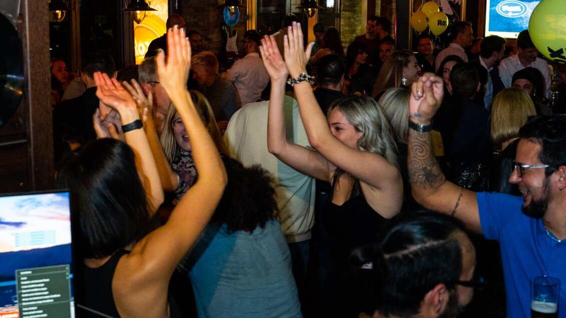 Gäste tanzen eng beieinander in einer vollen Bar, heben die Hände und lachen, im Hintergrund weitere Feiernde. | © ROX