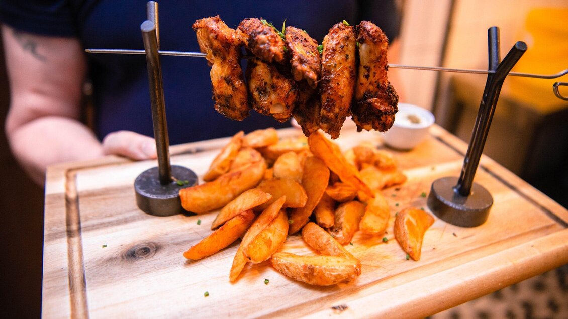 Ein Holzbrett mit knusprigen Hähnchenflügeln und frischen Kartoffelspalten. Die Speisen sind appetitlich angerichtet und werden auf serviert. | © ROX - Kyrillos Mikhail