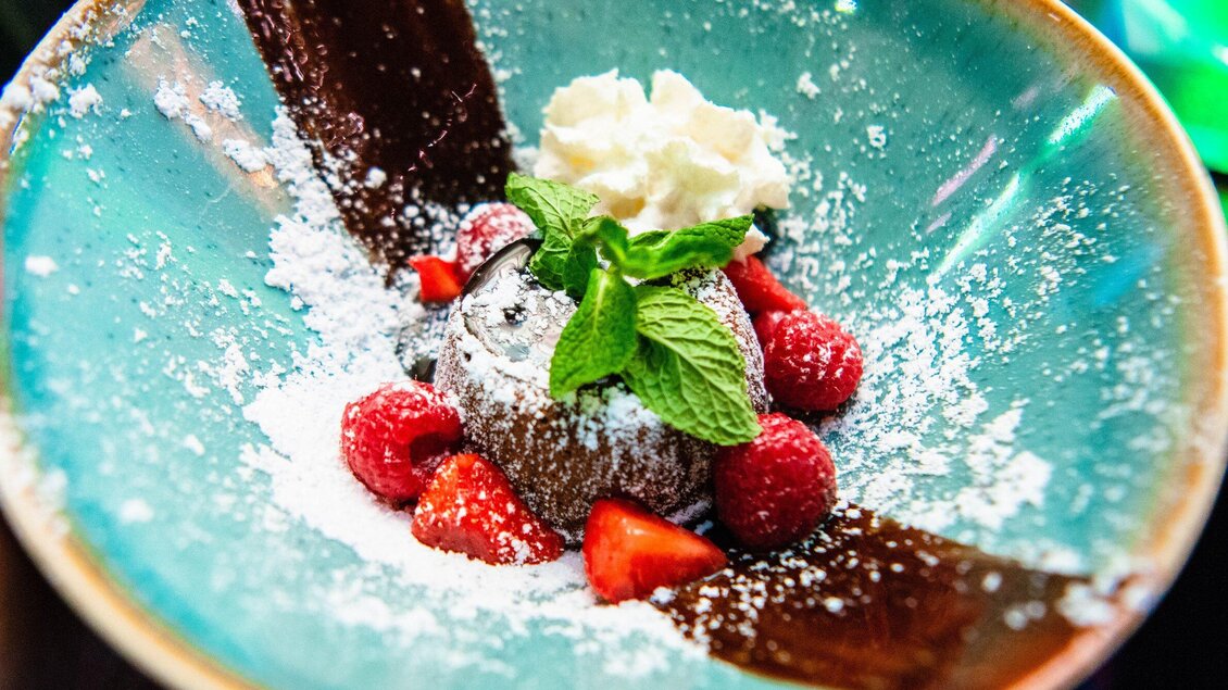 Ein leckerer Schokoladenkuchen auf einem blauen Teller, garniert mit frischen Beeren und einem Klecks Sahne. Zuckerstaub und Minzblätter verleihen dem Dessert das besondere Etwas. | © ROX - Kyrillos Mikhail
