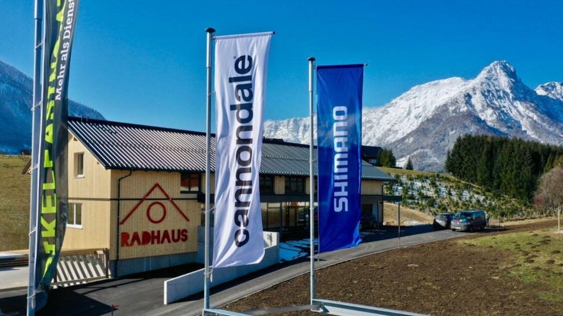 Ein moderner Fahrradladen mit dem Namen "RADHAUS" steht vor malerischen Bergen. Farbige Fahnen von verschiedenen Marken wehen im Wind. | © Radhaus, Bad Aussee, Luftaufnahme (c) Markus Raich