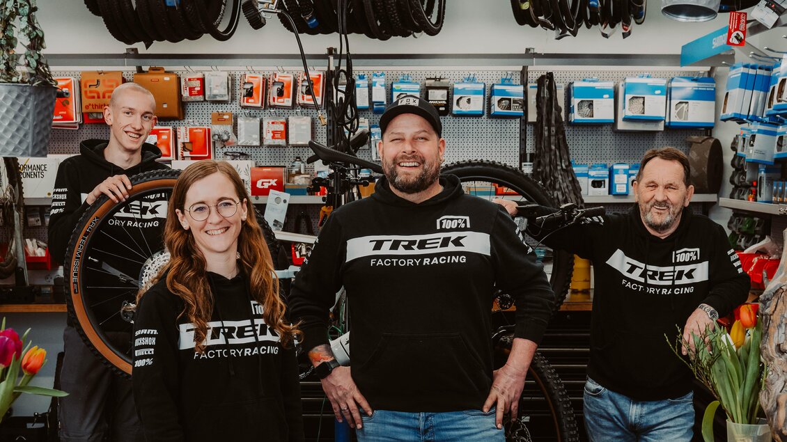 Ein Team von vier Personen in einem Fahrradgeschäft steht lächelnd hinter einem Verkaufstresen. Im Hintergrund sind Fahrradreifen und Zubehör sichtbar. | © Radwerkstatt Willibald