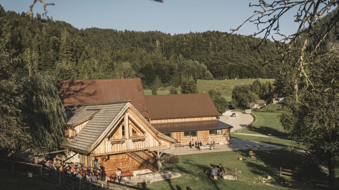 Ein rustikales Holzhaus umgeben von grünen Wiesen und Bäumen. Draußen sind Menschen versammelt und genießen die Natur. | © Stefan Leitner