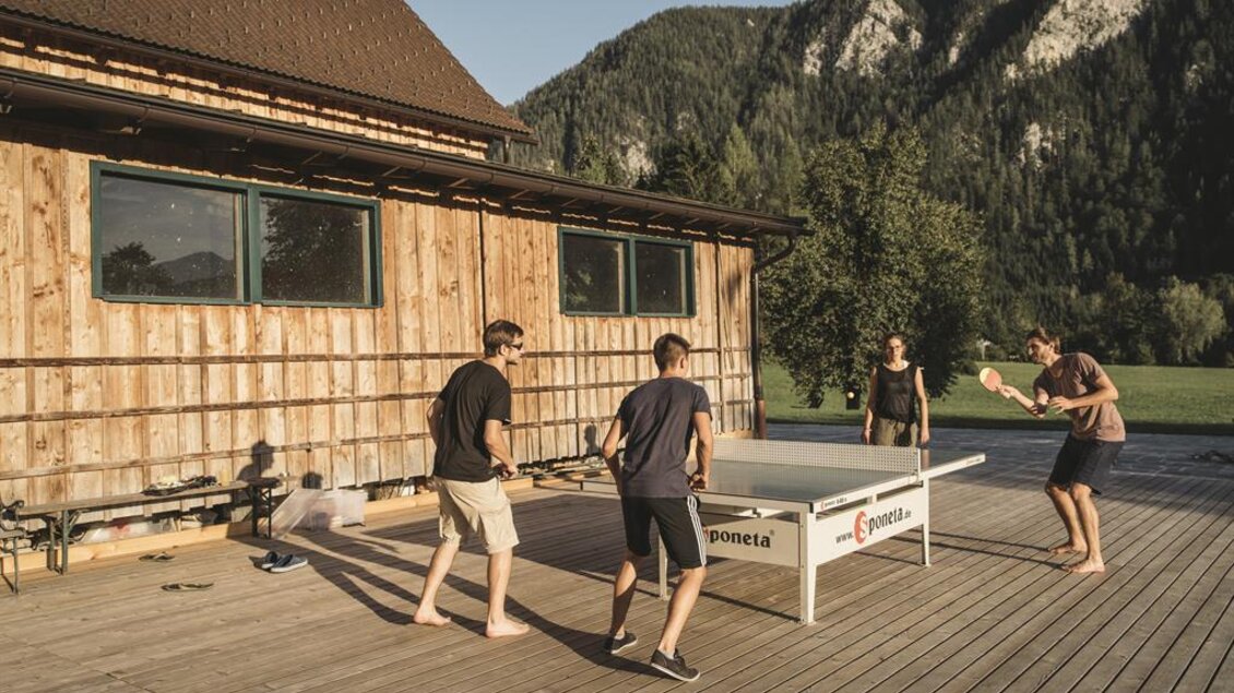 Eine Gruppe von vier Personen spielt Tischtennis auf einer Holzterrasse vor einem Holzhaus. Im Hintergrund sind grüne Berge zu sehen. | © Stefan Leitner