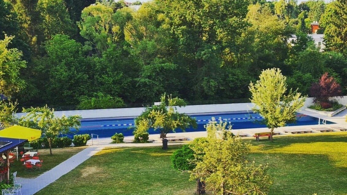 Das Ragnitzbad mit einem langen Schwimmbecken, umgeben von üppigem Grün. Auf der Wiese stehen einige Bäume und Sitzmöglichkeiten. | © Ragnitzbad