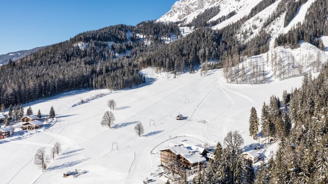 Eine malerische Schneelandschaft mit verschneiten Hügeln und vielen Tannenbäumen. Im Vordergrund stehen Chalets und eine Skipiste unter einem strahlend blauen Himmel.