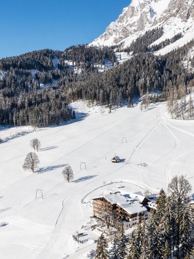 A picturesque snowy landscape with snow-covered hills and many fir trees. In the foreground, there are chalets and a ski slope under a bright blue sky.