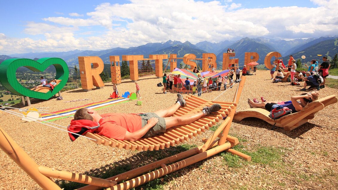 Eine Aussichtsplattform mit großen bunten Buchstaben von "RÜTISBERG" und einer Holzhängematte. Menschen entspannen und genießen die schöne Berglandschaft im Hintergrund. | © Hans-Peter Steiner