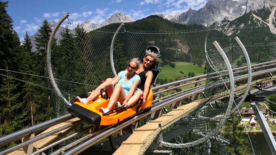 Zwei Personen fahren auf einer Sommerrodelbahn in den Bergen. Die Umgebung ist grün und die Berge sind klar sichtbar. | © Erlebnis Rittisberg