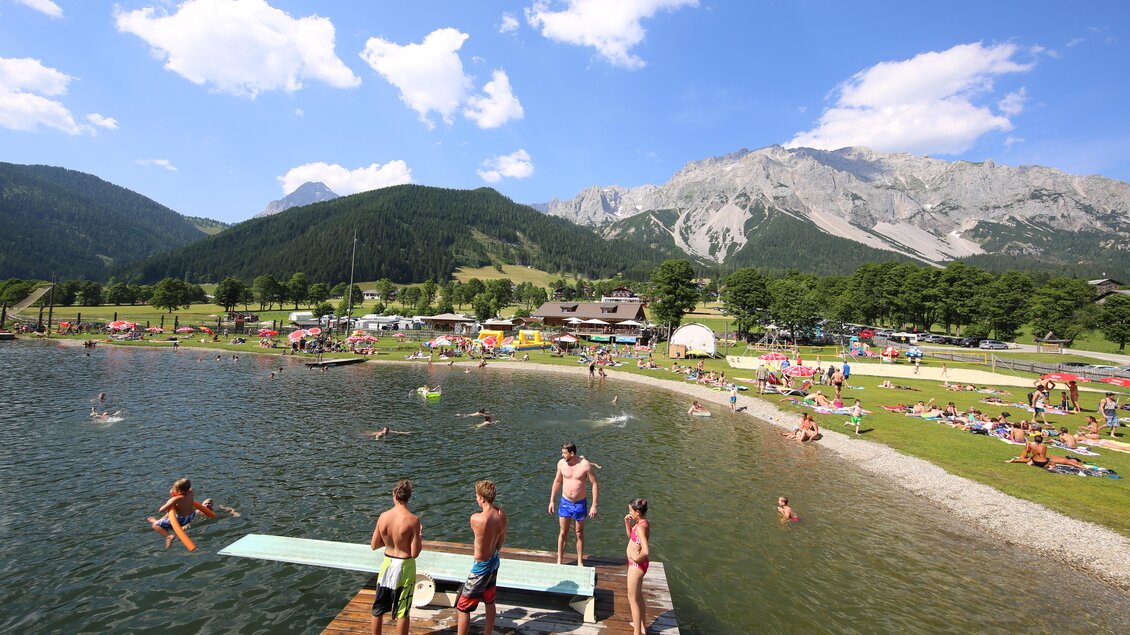 Ein schöner Badesee mit Menschen, die im Wasser schwimmen. Im Hintergrund sind Berge und eine grüne Landschaft zu sehen. | © Erlebnis Rittisberg