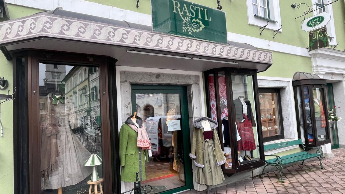 Ein Geschäft mit grüner Fassade und Schaufenstern, in denen traditionelle Kleidung ausgestellt ist. Vor dem Laden stehen Bänke und eine Blumendekoration. | © TVB Ausseerland Salzkammergut_ Nathalie Schröttenhamer