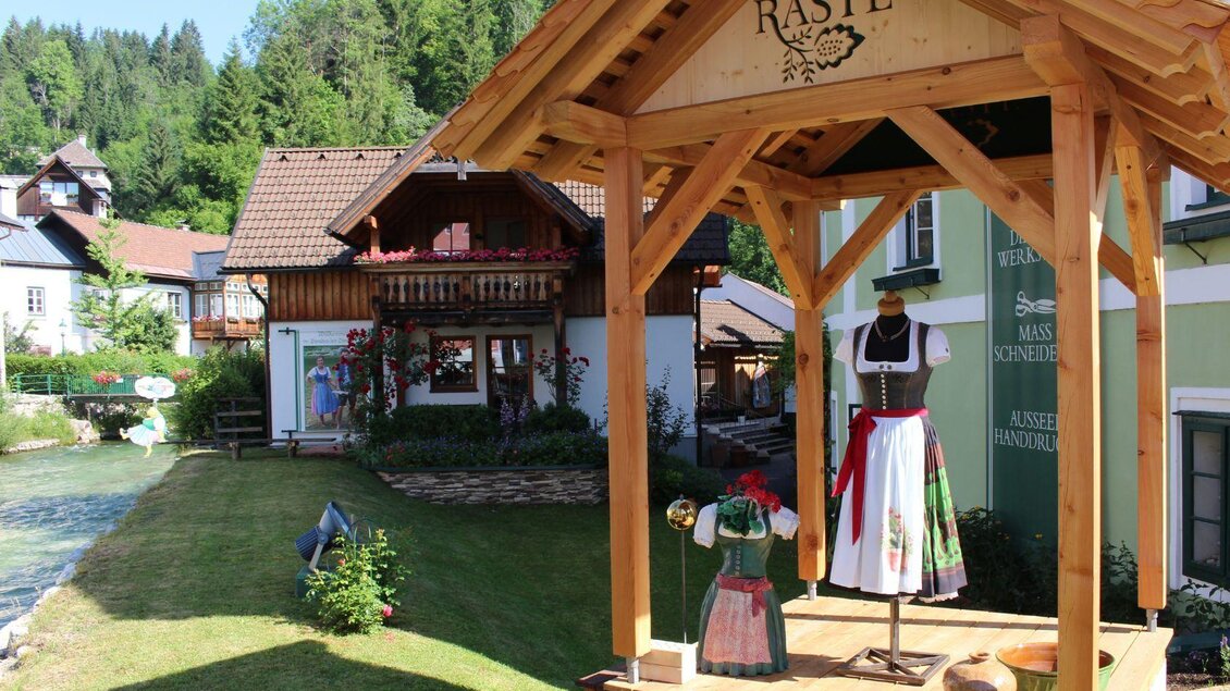 Ein traditioneller Holzstand mit Trachtenpuppen steht in einer malerischen Dorfansicht. Im Hintergrund sind grüne Hügel und liebevoll gestaltete Häuser zu sehen. | © TVB Ausseerland - Salzkammergut/Viola Lechner