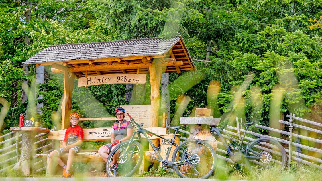 Ein schattiger Rastplatz in der Natur mit zwei Radfahrern, die auf einer Bank sitzen. Im Hintergrund sind Fahrräder und ein Schild mit der Höhenangabe 996 m zu sehen. | © Kräuterregion Wechselland / Oststeiermark Tourismus
