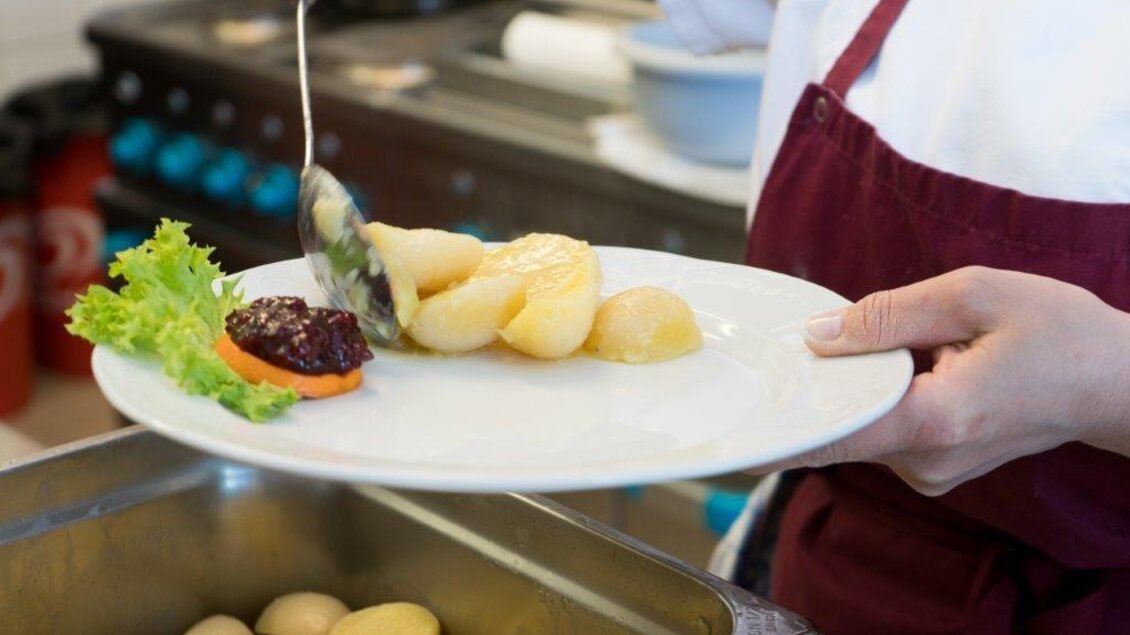 Ein Koch serviert Kartoffeln mit einem Klecks Sauce und einem Blatt Salat auf einem Teller. Im Hintergrund sind weitere Speisen sichtbar. | © Handler