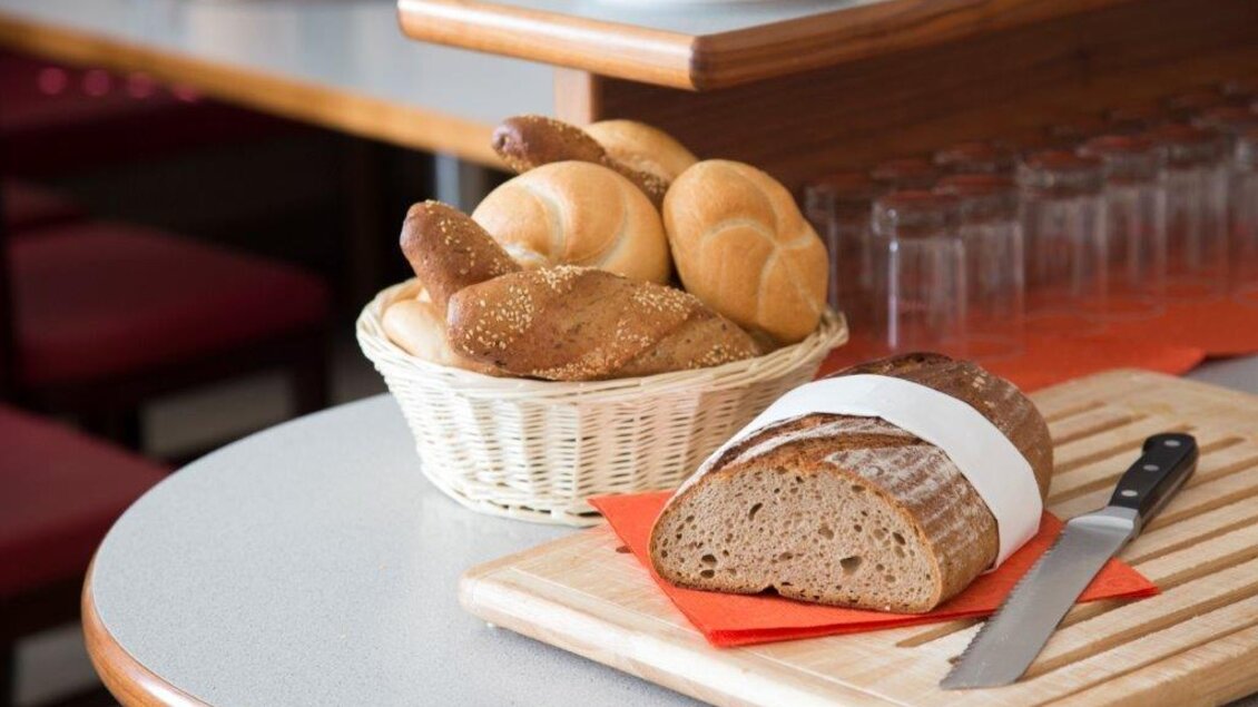 Ein Korb mit verschiedenen Brötchen und ein frisch geschnittenes Brot auf einem Holzbrett. Die Umgebung wirkt einladend und gemütlich. | © Handler