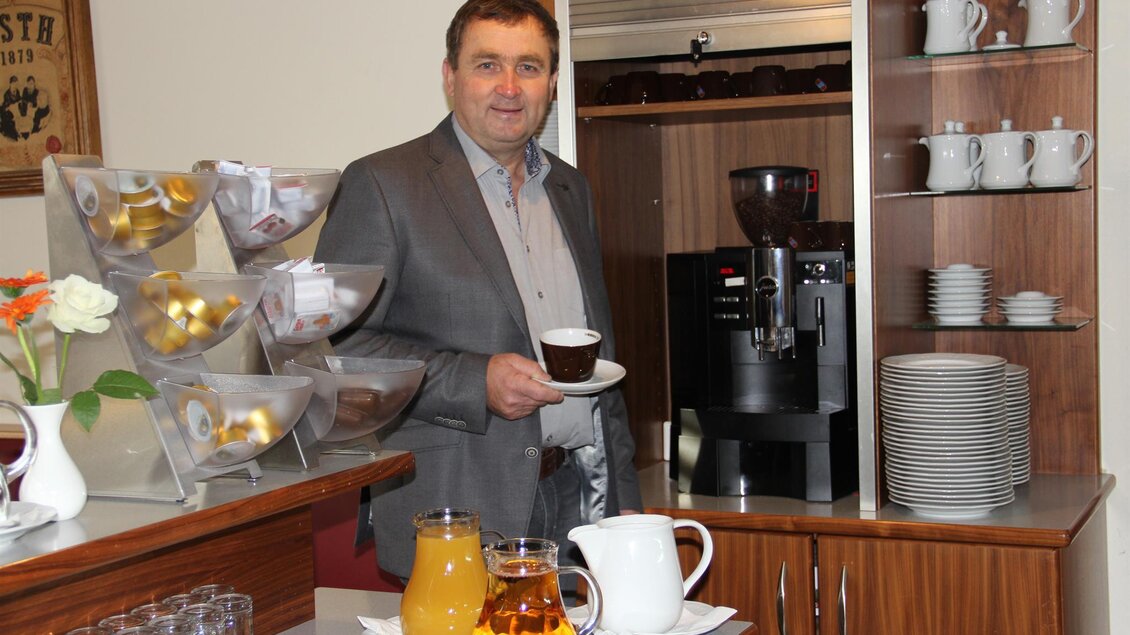 Ein Mann steht an einer Theke mit einer Tasse Kaffee in der Hand. Im Hintergrund sind eine Kaffeemaschine und verschiedene Getränke wie Saft und Tee zu sehen. | © Riedenbauer