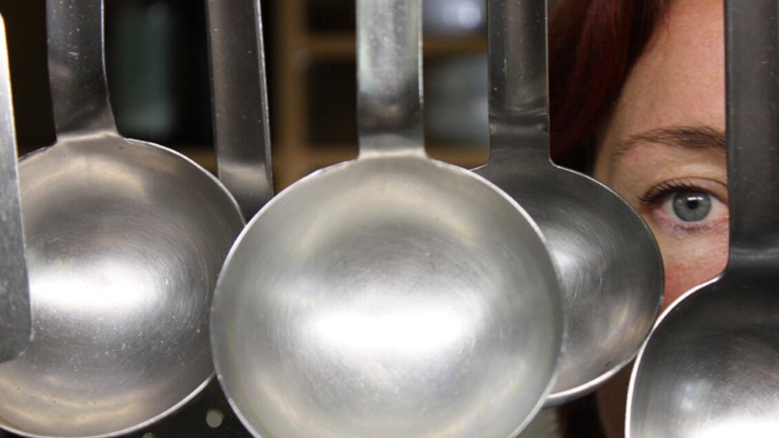 Eine Reihe von Metalllöffeln hängt in einer Küche. Im Hintergrund ist das Gesicht einer Person mit grünem Auge und rotem Haar zu sehen. | © Rauch-Hof