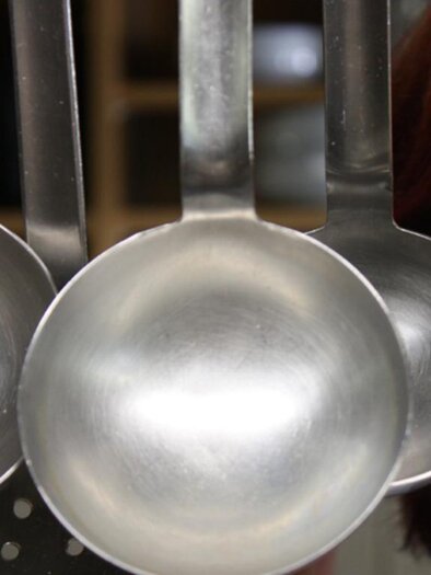 A series of metal spoons hangs in a kitchen. In the background, the face of a person with a green eye and red hair can be seen. | © Rauch-Hof