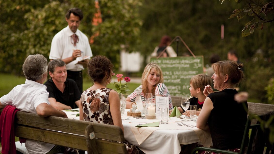 Eine Gruppe von sechs Personen sitzt an einem Tisch im Freien und genießt ein Essen. Im Hintergrund steht ein Kellner und ein grüner Garten umrahmt die Szene. | © Rauch-Hof