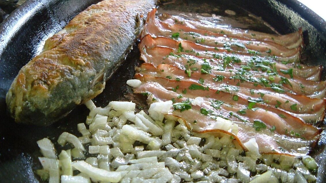 Ein gebratener Fisch liegt neben mehreren Scheiben Schinken in einer Pfanne. Darunter sind fein gehackte Zwiebeln sichtbar. | © Rauch-Hof